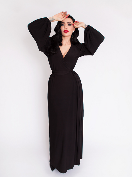 Micheline Pitt standing and posing with her hands positioned on her face while wearing the Black Widow Wrap Gown in Solid Black.