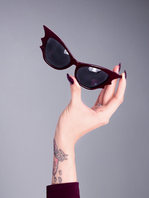 A product shot of a hand with oxblood colored nails holding the Vamp batwing sunglasses in oxblood.