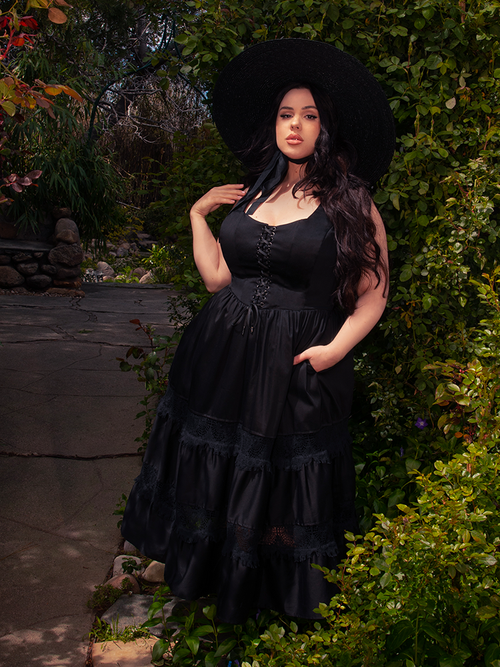 Amidst the blossoming garden, a brunette model basks in the sunlight, captivating all with her elegance in the enchanting Pickety Witch Dress in Black, a gothic masterpiece from La Femme en Noir.